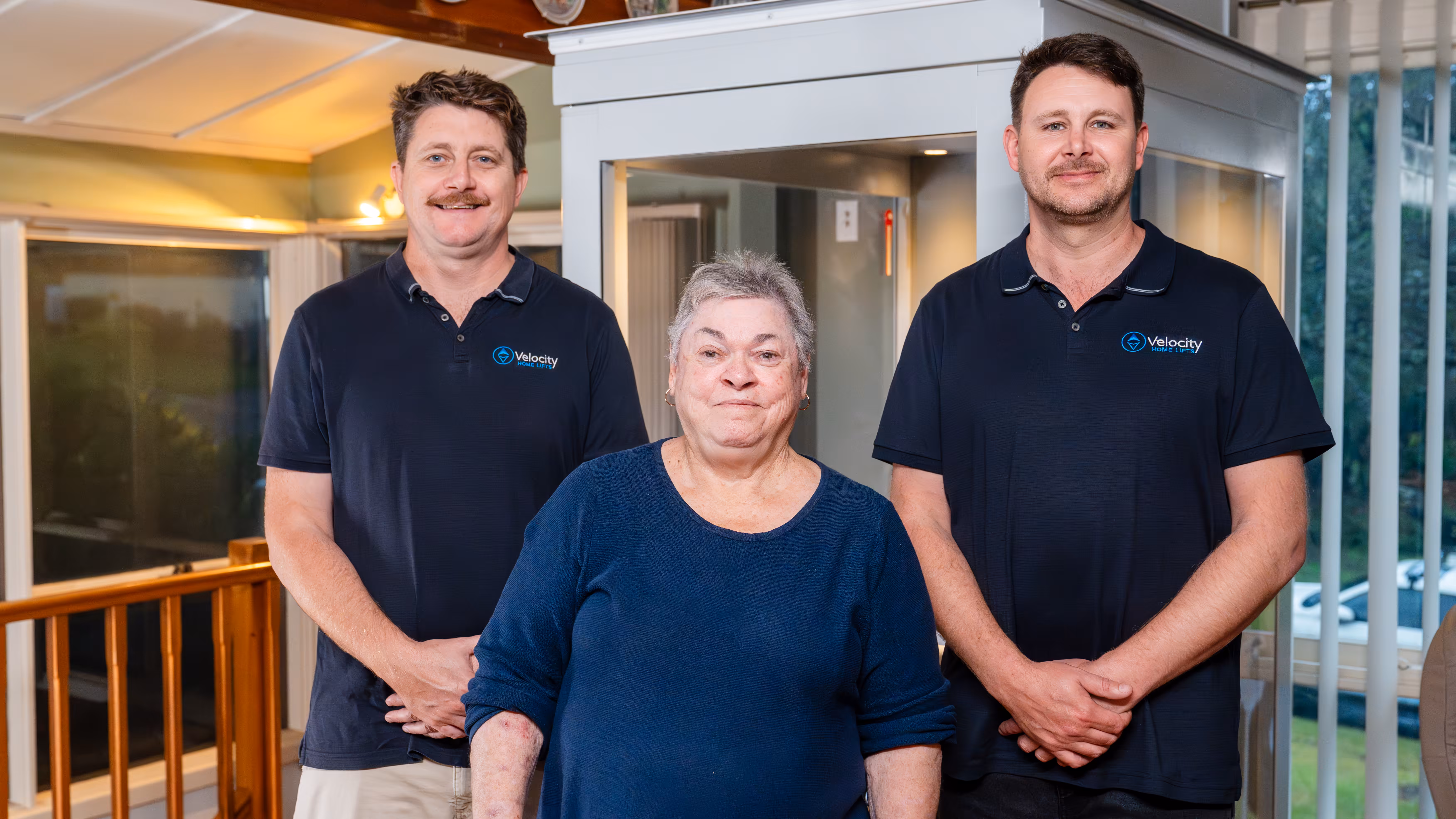 Two home lift owners standing with an elderly woman in the middle, all smiling in front of a lift