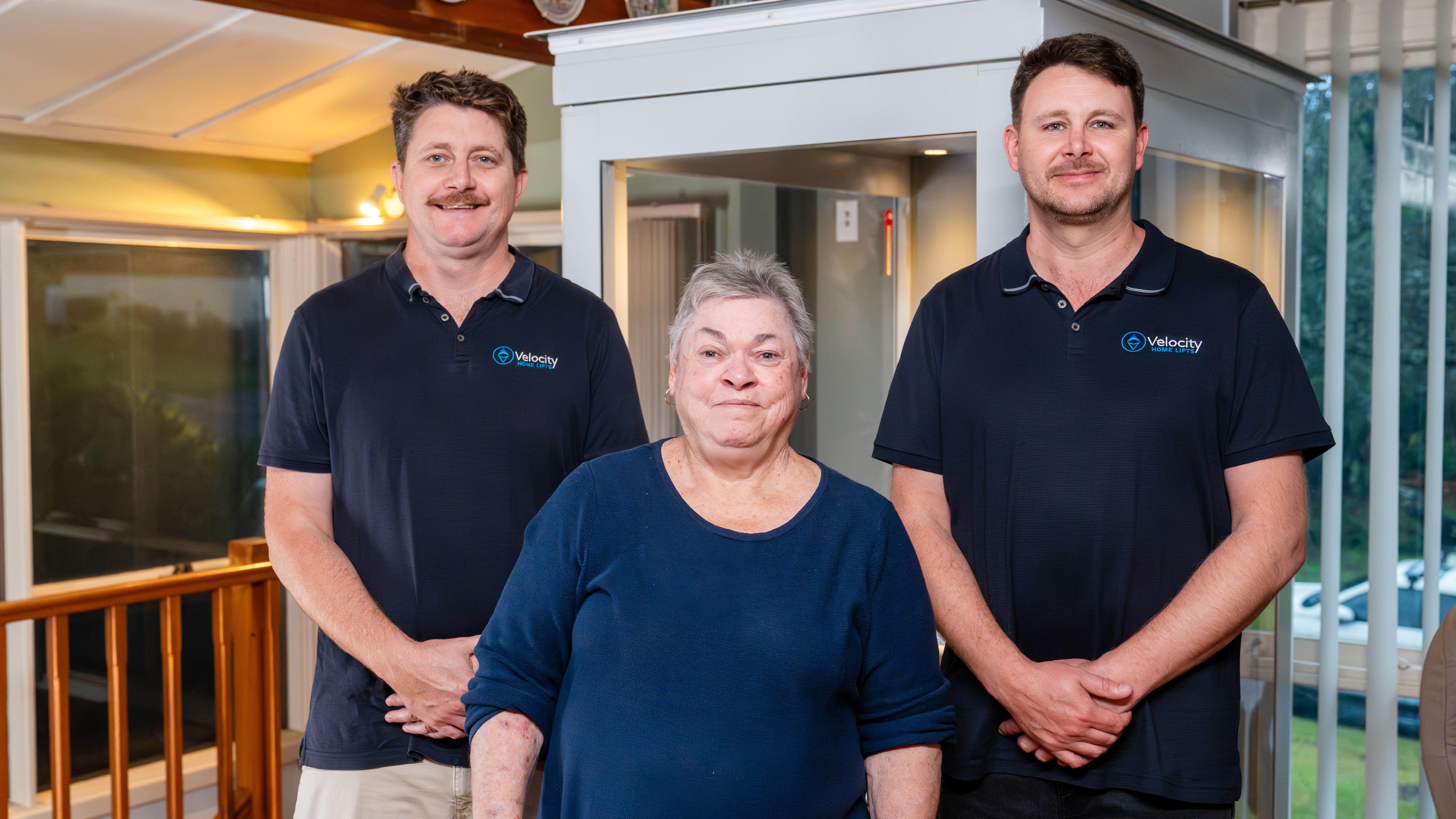 Two home lift owners standing with an elderly woman in the middle, all smiling in front of a lift